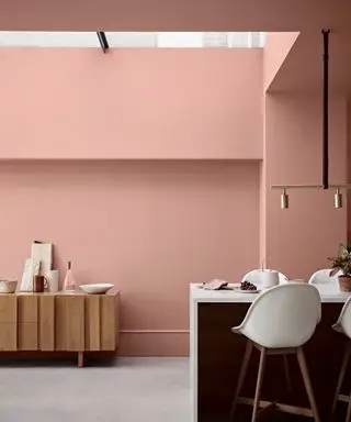 ピンクに塗られた地下室のキッチン、塗られた壁と天井、白とダークウッドのバーチェアのあるアイランドキッチン、グレーの床、背景に木製のサイドボード