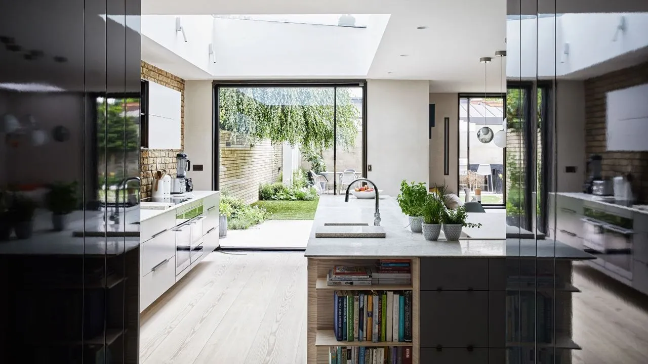 Una cucina moderna, luminosa e ariosa, con pavimento in legno, mobili bianchi e neri, un'isola con ripiani in marmo bianco e scaffalature integrate piene di libri di ricette, un'alzatina in mattoni a vista e grandi porte finestre su un rigoglioso giardino verde.' loading='lazy' title=