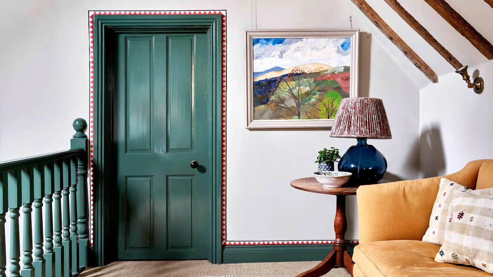 Un pianerottolo in cima a una scala con corrimano verdi e porta e rivestimento verdi abbinati. Dettagli rossi dipinti attorno al rivestimento. Un divano giallo sul lato destro accanto a un tavolino rotondo in legno con sopra una lampada.' loading='lazy' title=