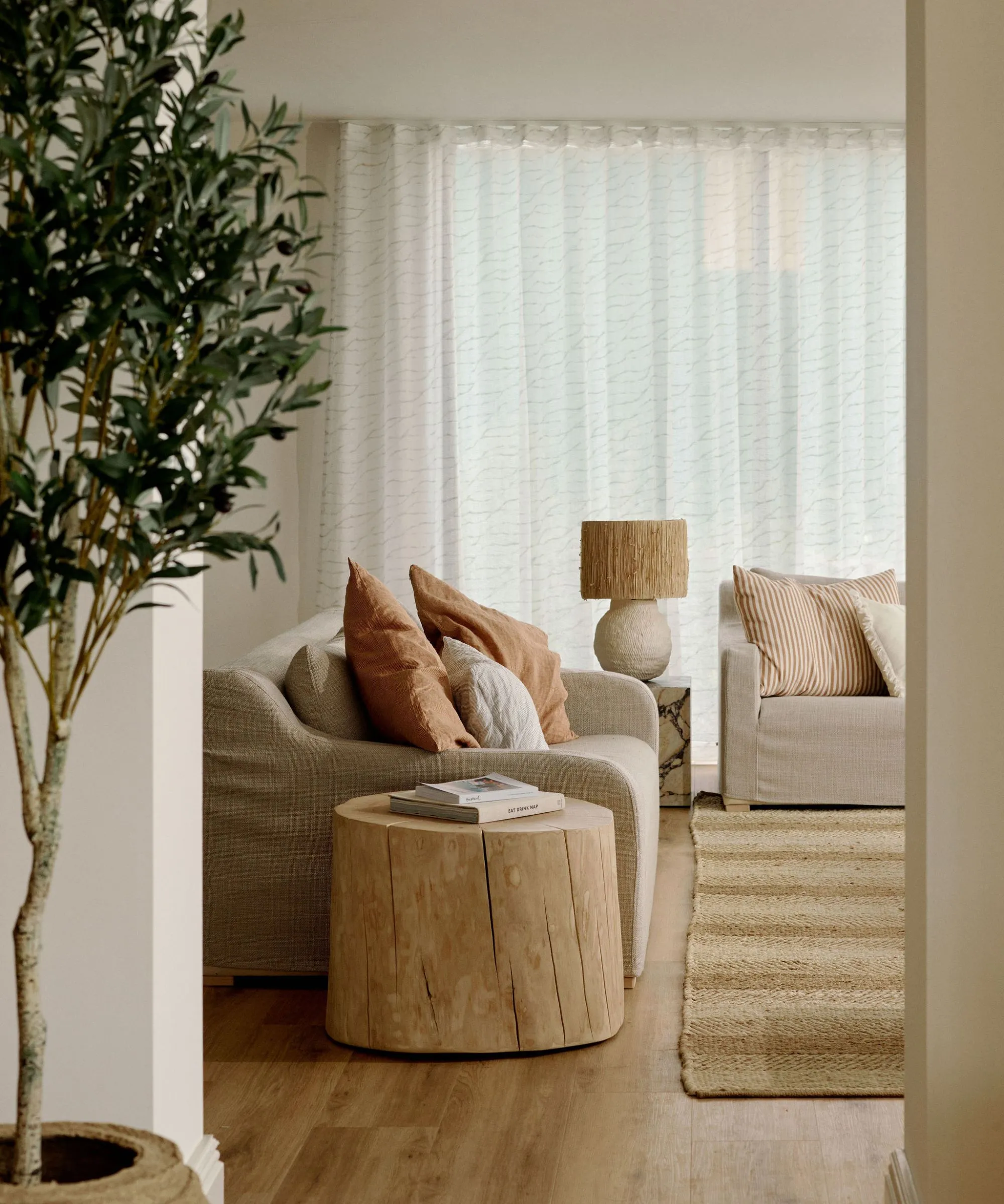 soggiorno neutro, caldo e minimalista, con un tappeto di iuta, due divani ricoperti in lino, un tavolino rustico in legno e un ulivo' loading='lazy' title=