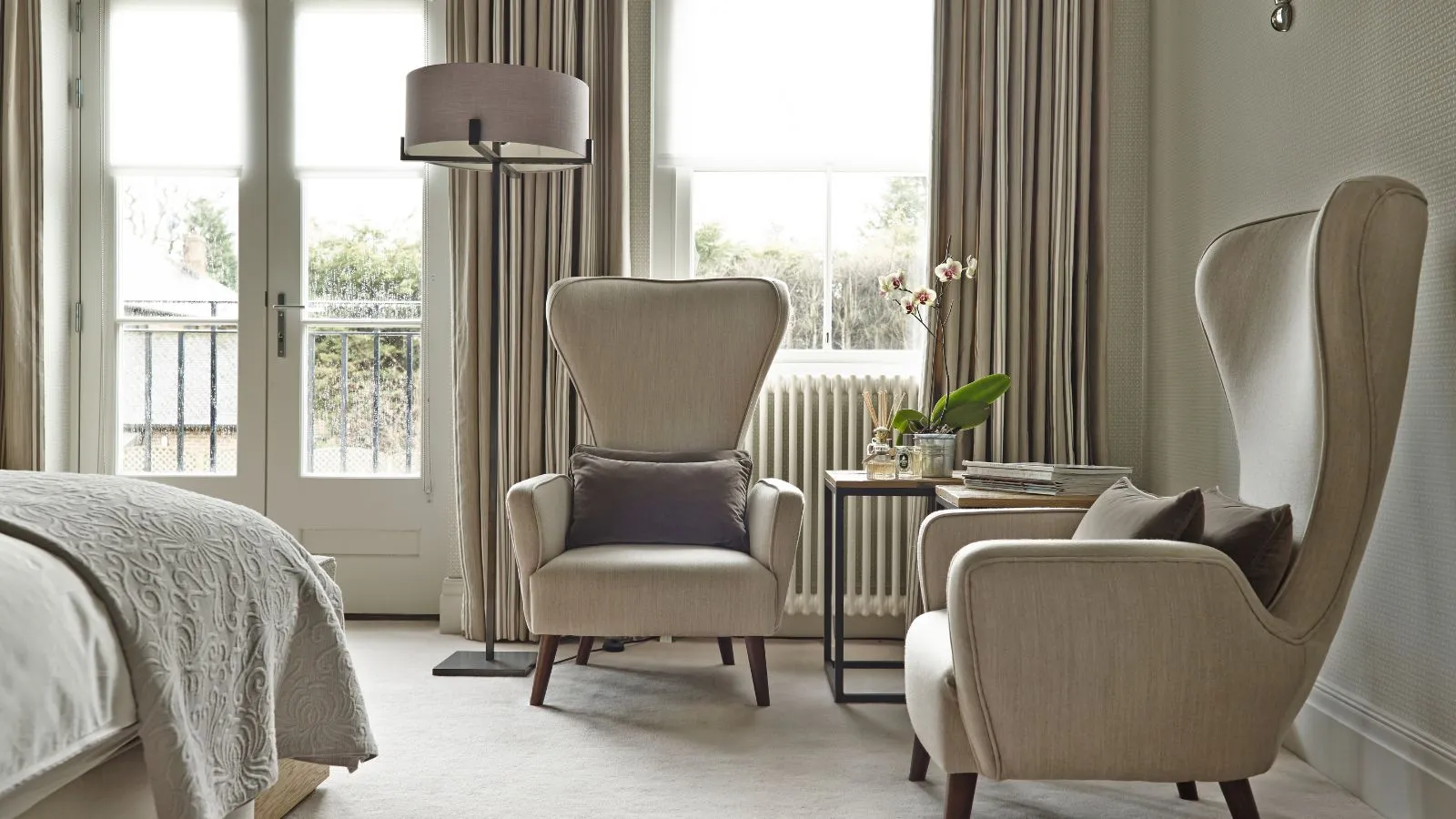 Ein Teppich -Sahne -Schlafzimmer mit großen gepolsterten Stühlen mit braunen Kissen, die große Fenster mit einer hohen Lampe und einem Tisch mit einer Blume und einem Diffusor befinden. In der linken Hand ist die Ecke eines Bettes zu sehen.' loading='lazy' title=