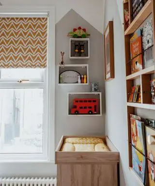 グレーの抽象的なデザインのペイントされた子供用ベッドルーム、おもちゃや装飾アイテムを備えた壁掛け木製棚、壁掛け木製本棚、黄色の模様のブラインド、白塗装のベビーベッド